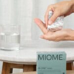 Person holding a Miome IB-ONE probiotic supplement capsule above a glass of water on a small marble table.
