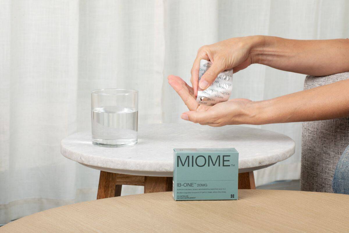 Person holding a Miome IB-ONE probiotic supplement capsule above a glass of water on a small marble table.