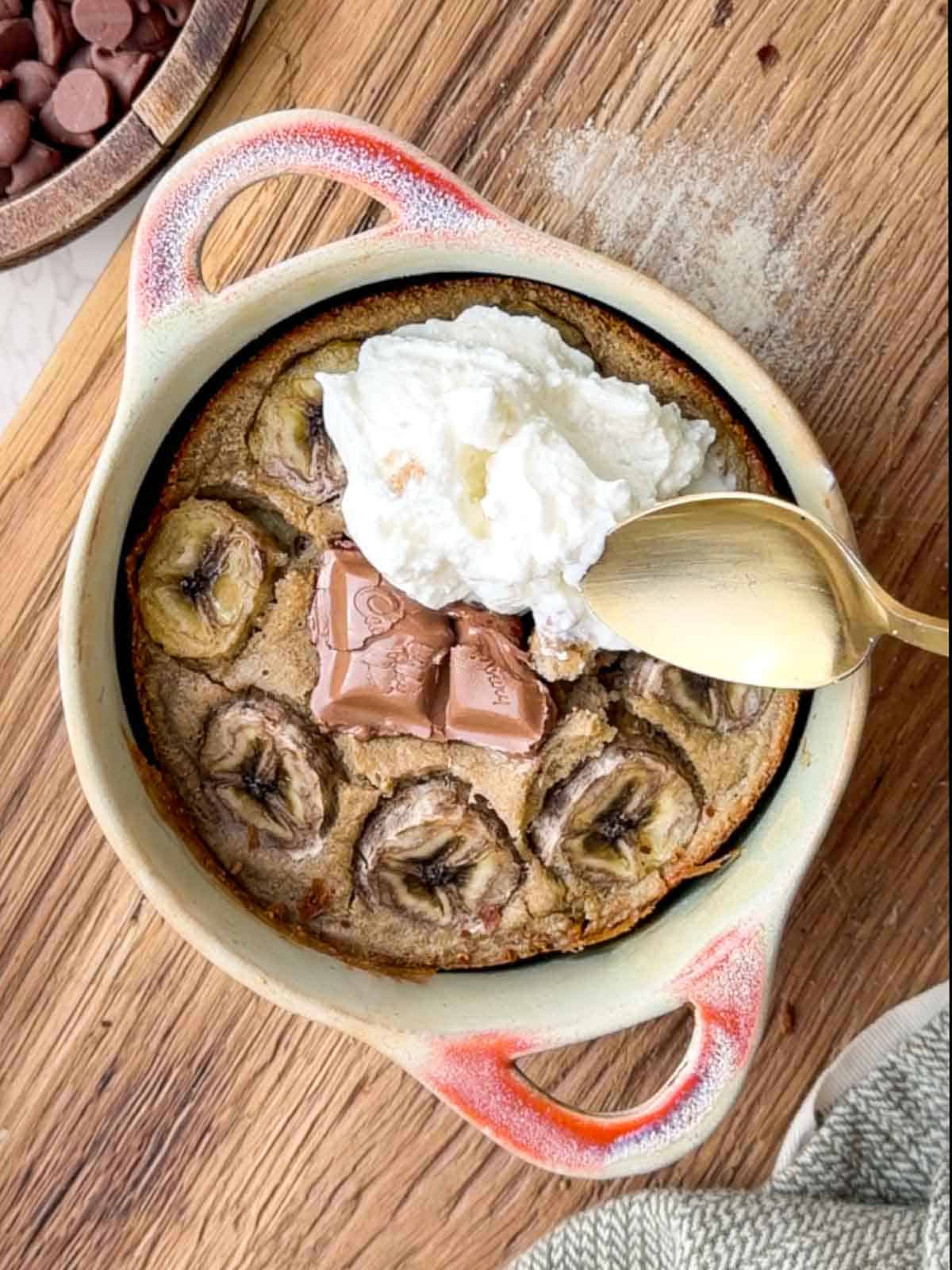 Banana bread baked oats in a small ceramic dish, topped with sliced banana, melted chocolate pieces, and a dollop of yogurt, served with a spoon on a wooden board.