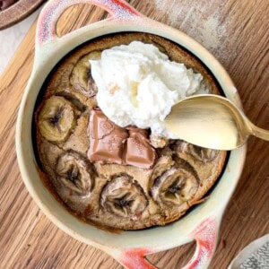 Banana bread baked oats in a small ceramic dish, topped with sliced banana, melted chocolate pieces, and a dollop of yogurt, served with a spoon on a wooden board.