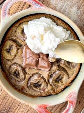 Banana bread baked oats in a small ceramic dish, topped with sliced banana, melted chocolate pieces, and a dollop of yogurt, served with a spoon on a wooden board.