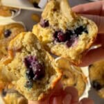 Banana cottage cheese muffins split in half showing a soft, fluffy interior with juicy blueberries, held by hand over a tray of freshly baked muffins in paper liners.