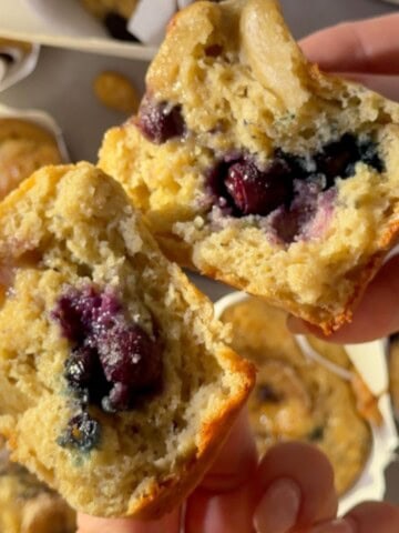 Banana cottage cheese muffins split in half showing a soft, fluffy interior with juicy blueberries, held by hand over a tray of freshly baked muffins in paper liners.