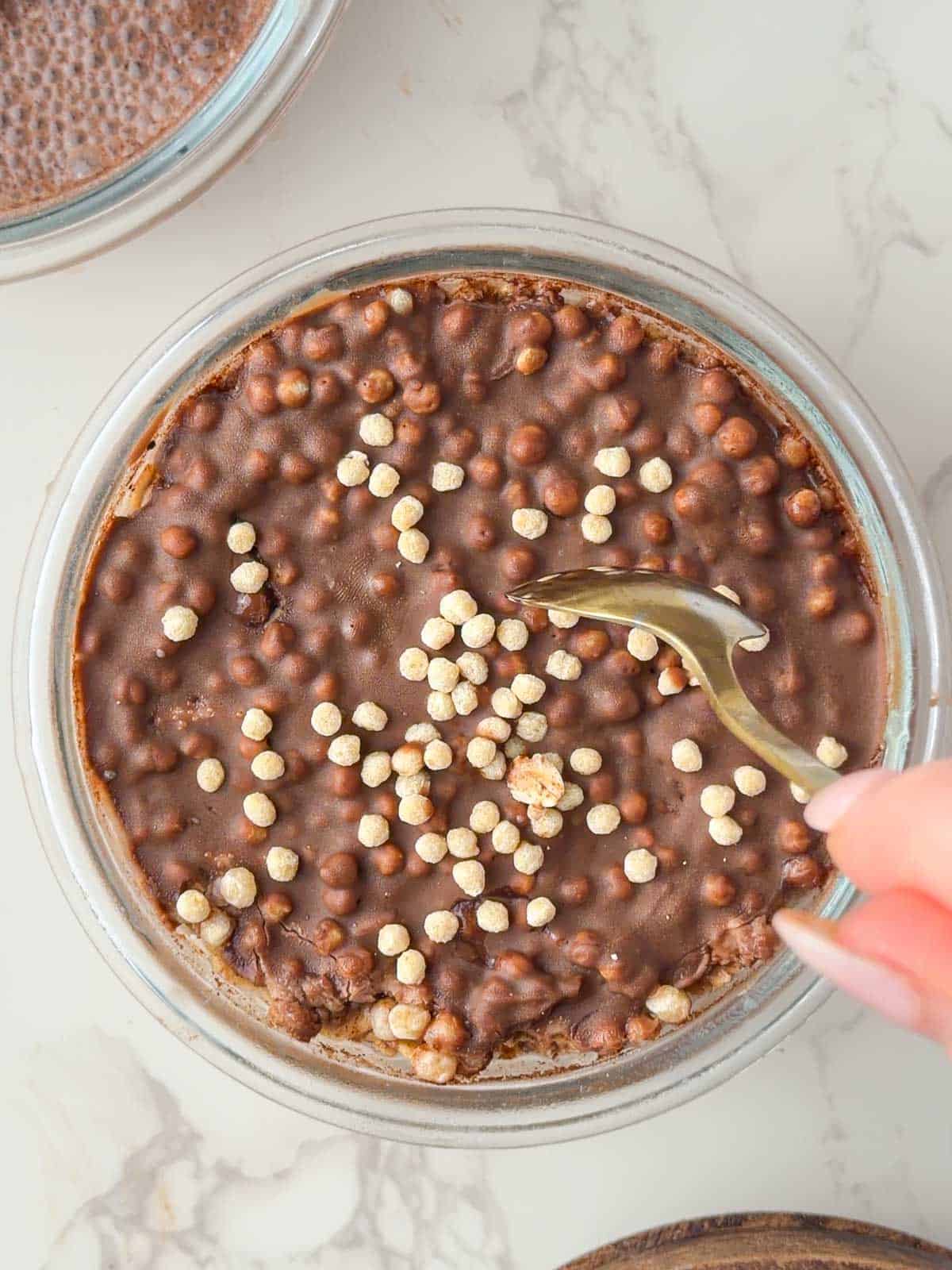 Chocolate protein overnight oats topped with a smooth, crackable chocolate shell broken with a spoon in a glass bowl
