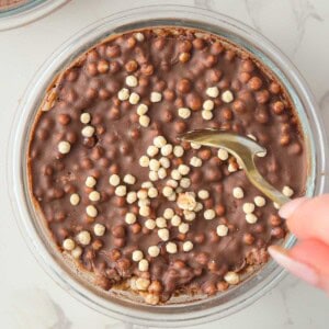 Chocolate protein overnight oats topped with a smooth, crackable chocolate shell broken with a spoon in a glass bowl