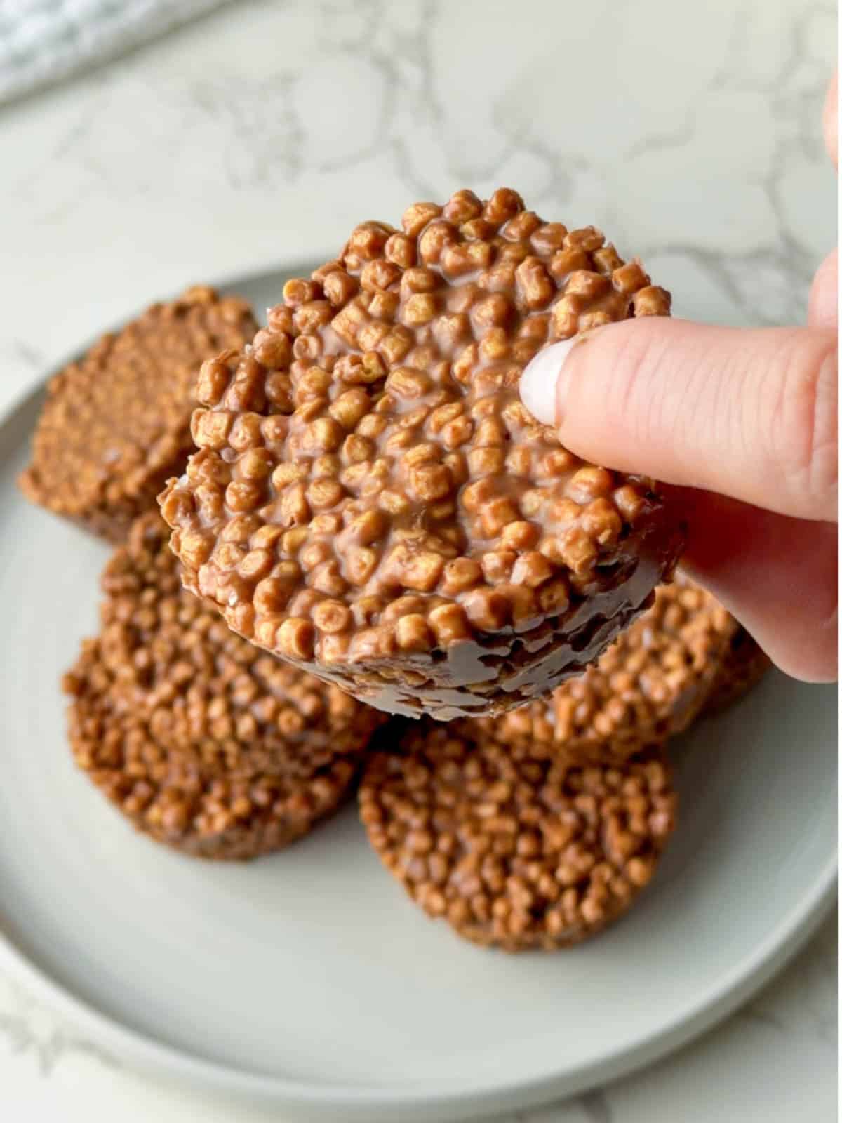 High protein chocolate crispy cakes made with soy or pea protein crisps and coated in chocolate, stacked on a white plate with one being held up to show the crunchy texture. These no bake chocolate crispy cakes are kid friendly, easy to make, and perfect for a high protein snack or dessert.