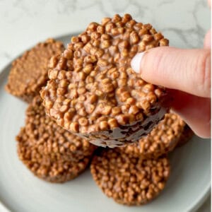 High protein chocolate crispy cakes made with soy or pea protein crisps and coated in chocolate, stacked on a white plate with one being held up to show the crunchy texture. These no bake chocolate crispy cakes are kid friendly, easy to make, and perfect for a high protein snack or dessert.