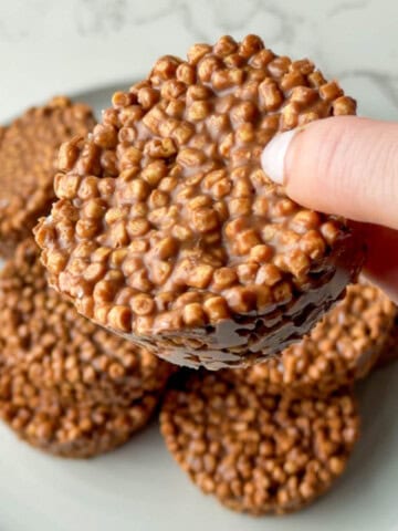 High protein chocolate crispy cakes made with soy or pea protein crisps and coated in chocolate, stacked on a white plate with one being held up to show the crunchy texture. These no bake chocolate crispy cakes are kid friendly, easy to make, and perfect for a high protein snack or dessert.