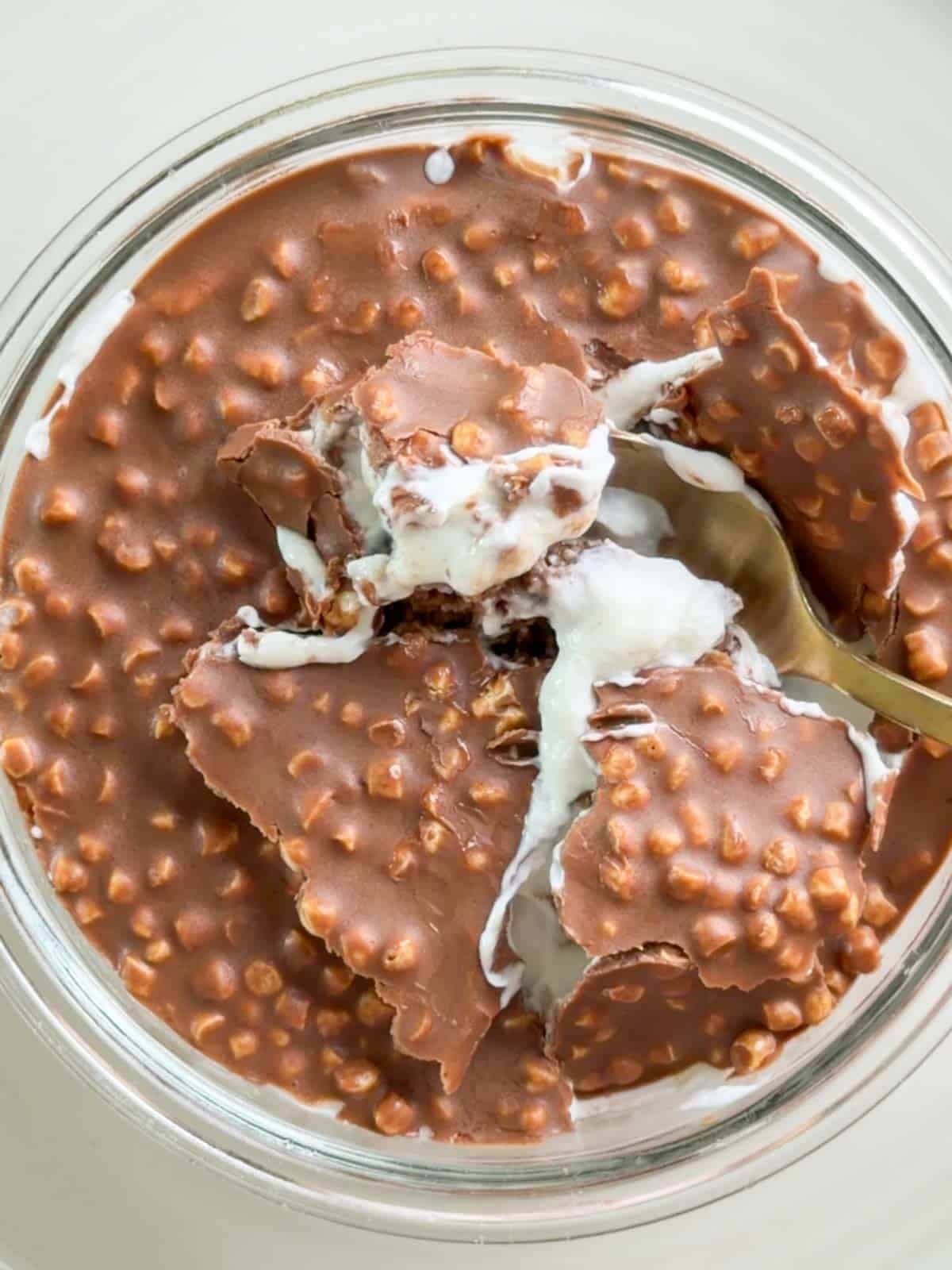 Close-up of creamy Maltesers Overnight Weetabix in a glass dish with a thick crunchy chocolate topping made with crisp cereal pieces. A spoon scoops through the cracked chocolate layer into the creamy overnight Weetabix underneath.