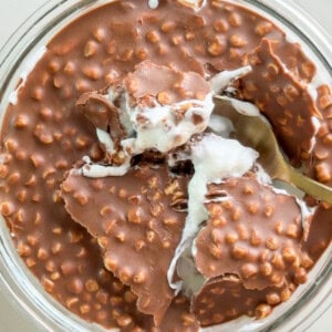 Close-up of creamy Maltesers Overnight Weetabix in a glass dish with a thick crunchy chocolate topping made with crisp cereal pieces. A spoon scoops through the cracked chocolate layer into the creamy overnight Weetabix underneath.