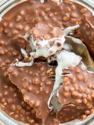 Close-up of creamy Maltesers Overnight Weetabix in a glass dish with a thick crunchy chocolate topping made with crisp cereal pieces. A spoon scoops through the cracked chocolate layer into the creamy overnight Weetabix underneath.