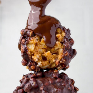 Chocolate peanut butter protein balls stacked with a gooey chocolate drizzle pouring over the top, showing the crispy rice texture inside. These no-bake peanut butter rice krispie balls are coated in chocolate and made with simple high-protein ingredients.