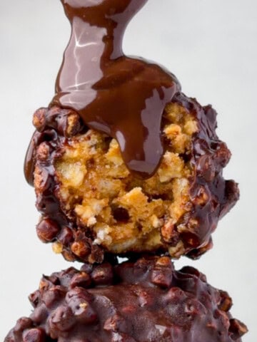 Chocolate peanut butter protein balls stacked with a gooey chocolate drizzle pouring over the top, showing the crispy rice texture inside. These no-bake peanut butter rice krispie balls are coated in chocolate and made with simple high-protein ingredients.