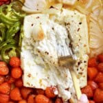 Spicy feta pasta ingredients roasting in a baking dish with a block of feta cheese, cherry tomatoes, jalapeño slices, roasted red peppers, onion, and garlic. A fork presses into the creamy baked feta before mixing.
