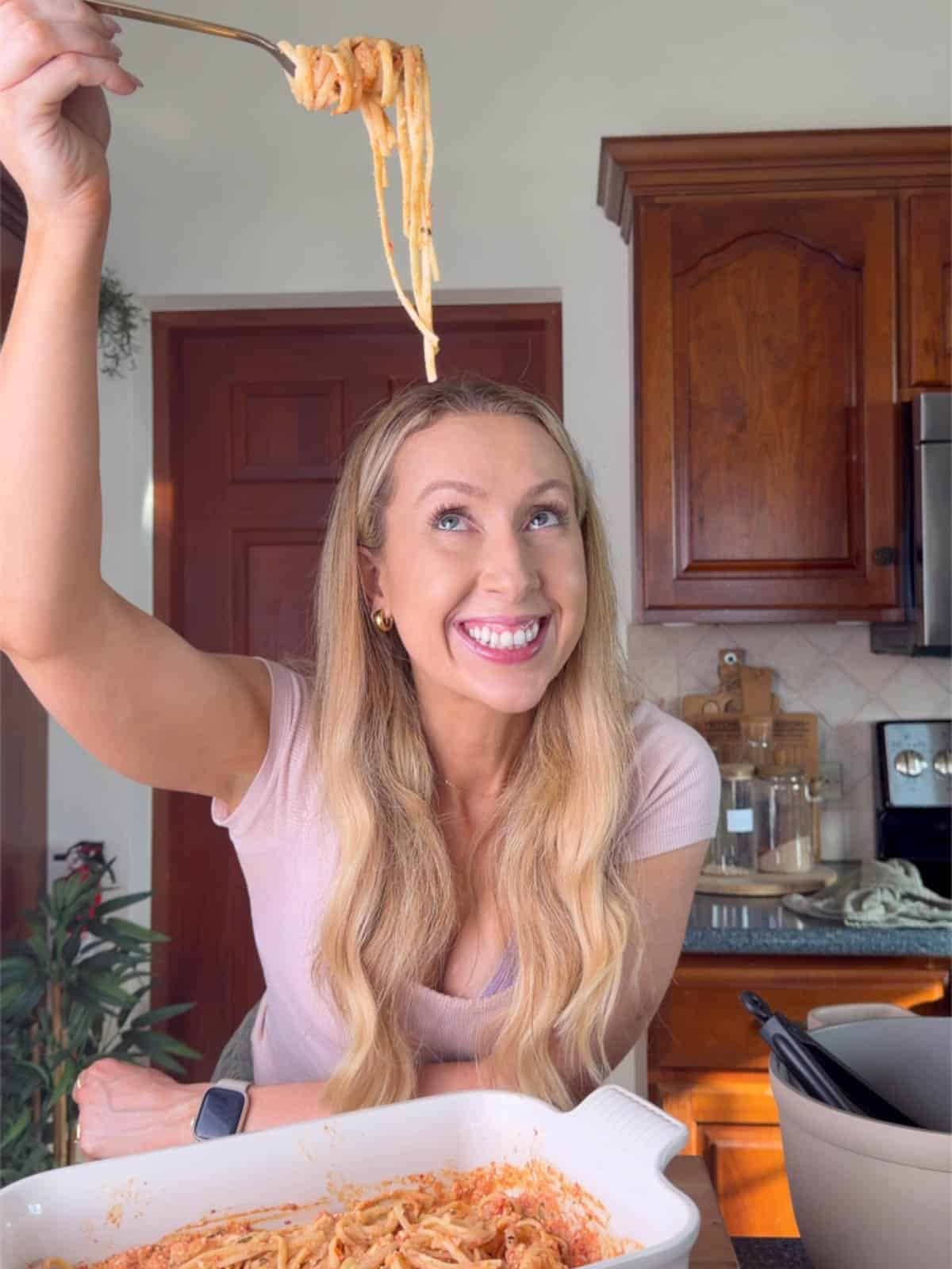 Woman (maria lucey dietitian) holding up a forkful of spicy feta pasta in a kitchen, smiling as she lifts creamy tomato feta spaghetti from a casserole dish.