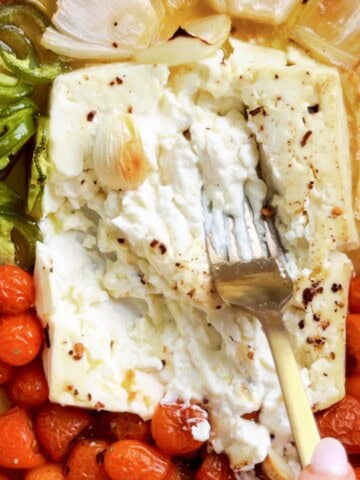 Spicy feta pasta ingredients roasting in a baking dish with a block of feta cheese, cherry tomatoes, jalapeño slices, roasted red peppers, onion, and garlic. A fork presses into the creamy baked feta before mixing.