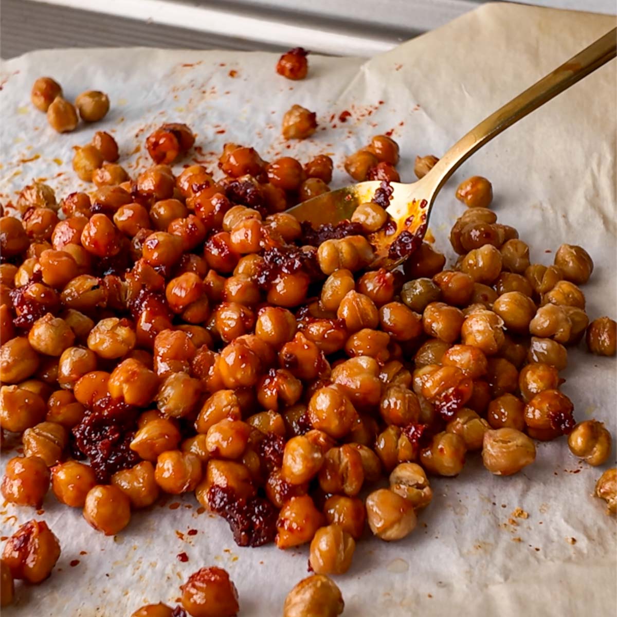 Spicy roasted chickpeas with harissa served on parchment paper with a gold spoon. The crispy chickpeas are coated in smoky harissa paste, giving them a rich red color and bold flavor.