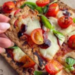 Close-up of a crispy flatbread Margherita pizza topped with melted fresh mozzarella, roasted cherry tomatoes, fresh basil leaves, and a drizzle of balsamic glaze. A hand holds up a slice, showing the thin, golden-brown crust and bubbling cheese.