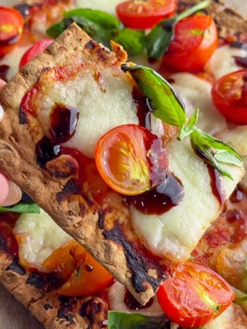 Close-up of a crispy flatbread Margherita pizza topped with melted fresh mozzarella, roasted cherry tomatoes, fresh basil leaves, and a drizzle of balsamic glaze. A hand holds up a slice, showing the thin, golden-brown crust and bubbling cheese.