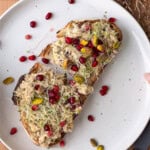 Two slices of toasted sourdough topped with creamy sardine salad, pomegranate arils, pistachios, and sprouts on a white plate.