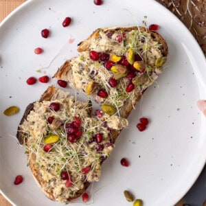 Two slices of toasted sourdough topped with creamy sardine salad, pomegranate arils, pistachios, and sprouts on a white plate.