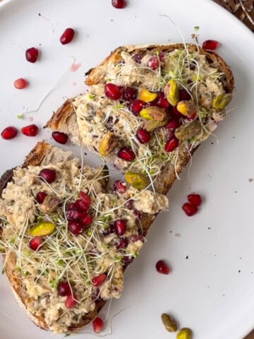 Two slices of toasted sourdough topped with creamy sardine salad, pomegranate arils, pistachios, and sprouts on a white plate.
