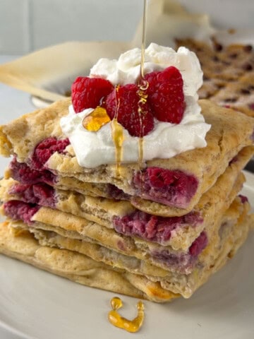 Stack of sheet pan protein pancakes filled with raspberries and topped with whipped yogurt, fresh raspberries, and a drizzle of honey.