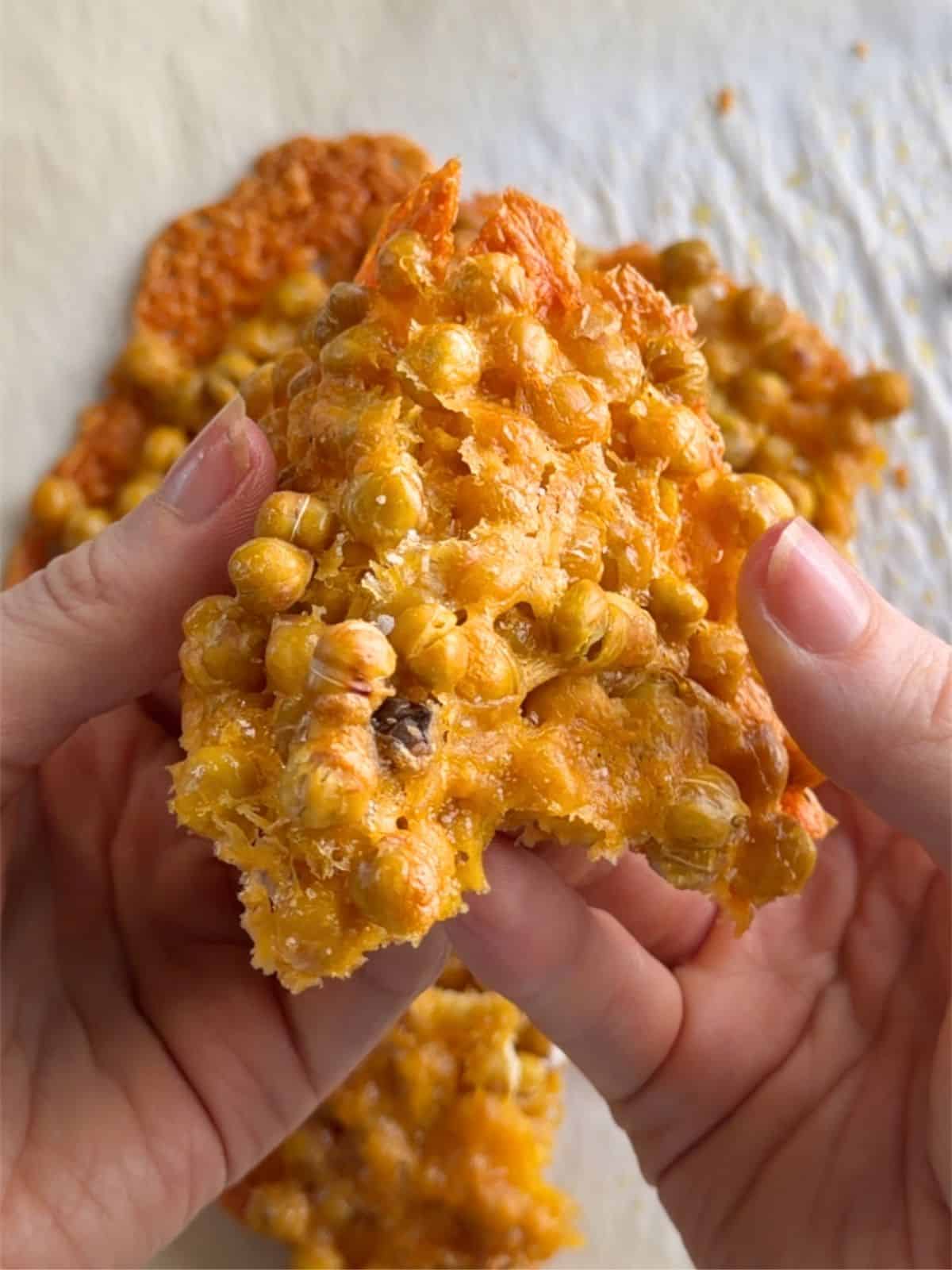 Cheesy Chickpea Chips held in hands, showing a crispy, golden baked chickpea and cheese cracker with a crunchy texture.