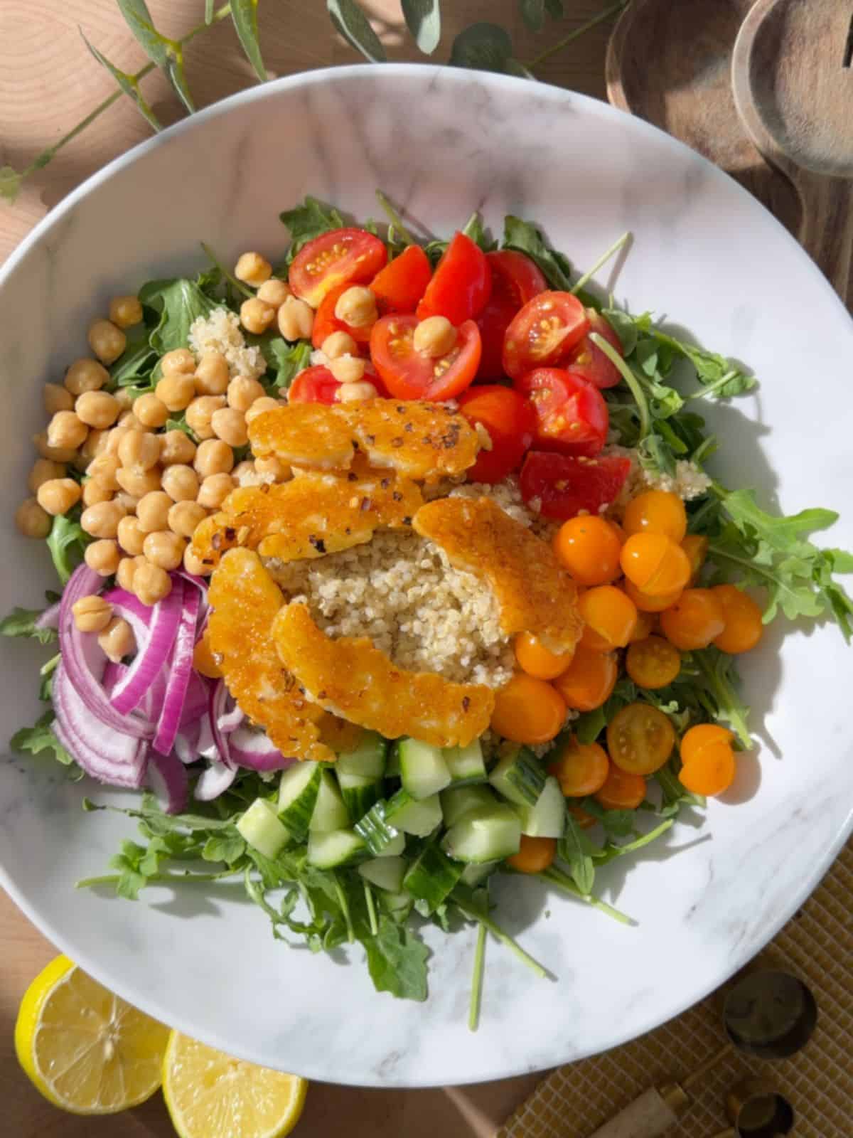 Chickpea and halloumi salad with crispy golden halloumi, chickpeas, quinoa, arugula, cucumber, red onion and colorful cherry tomatoes in a white bowl.
