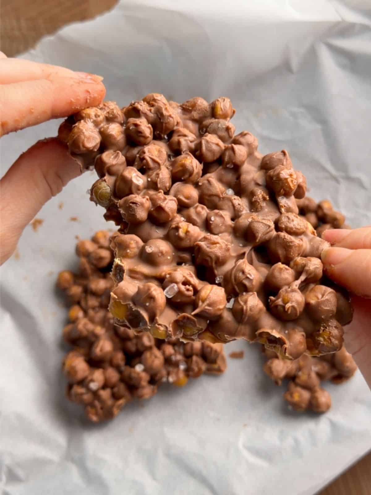 Hands breaking apart a piece of chocolate chickpea bark made with crispy roasted chickpeas, melted chocolate and a sprinkle of flaky sea salt.