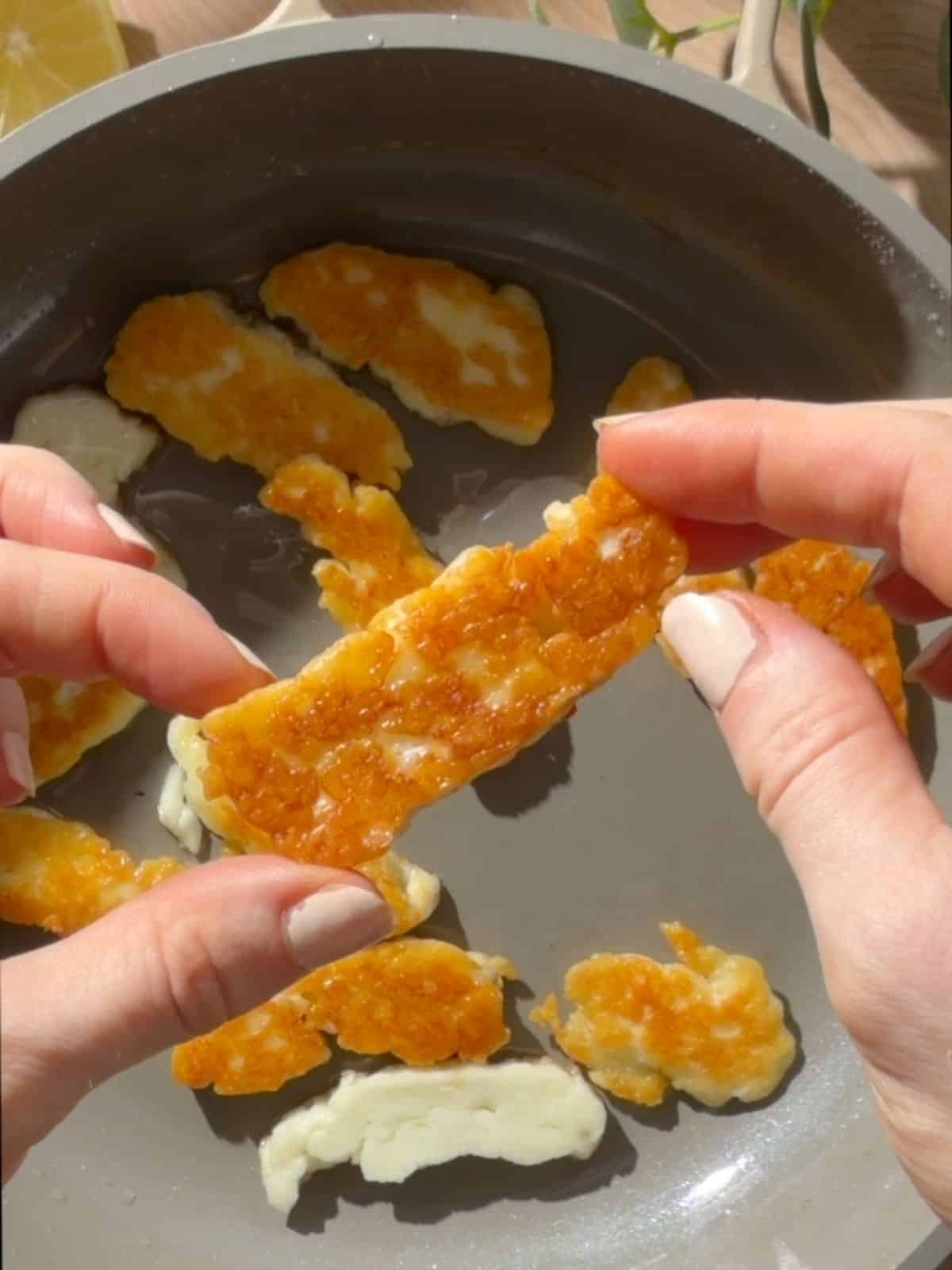 A slice of halloumi in between someones hands showing its golden color with a pan of more halloumi in the background.