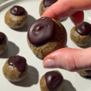 Hand holding a nut free protein ball topped with dark chocolate, with more chocolate-topped protein balls on a plate.