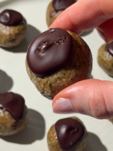 Hand holding a nut free protein ball topped with dark chocolate, with more chocolate-topped protein balls on a plate.