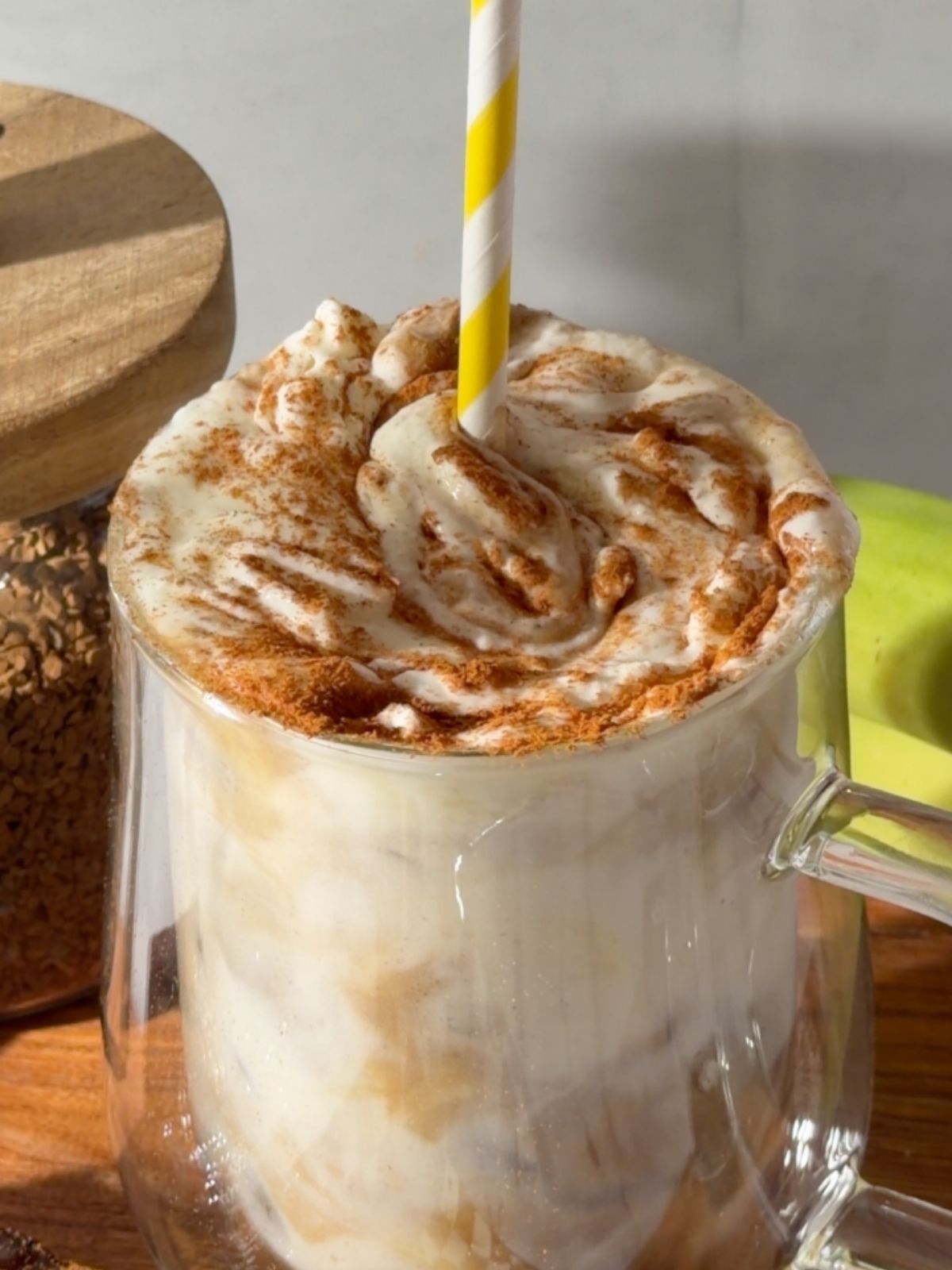 Creamy banana bread latte topped with whipped cream and a sprinkle of cinnamon, served in a clear glass mug with a yellow-and-white striped straw.