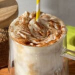 Creamy banana bread latte topped with whipped cream and a sprinkle of cinnamon, served in a clear glass mug with a yellow-and-white striped straw.