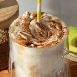 Creamy banana bread latte topped with whipped cream and a sprinkle of cinnamon, served in a clear glass mug with a yellow-and-white striped straw.