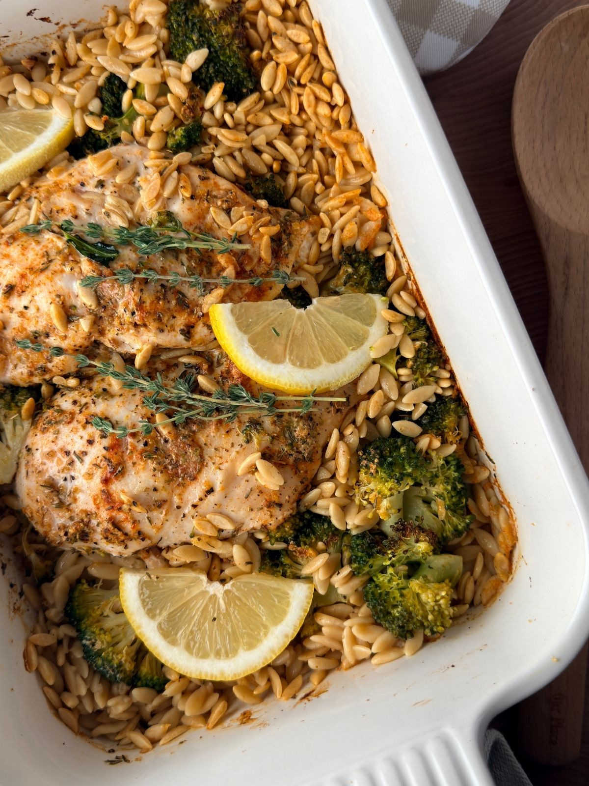 Lemon chicken orzo pasta bake with broccoli, fresh thyme and lemon wedges in a white baking dish.