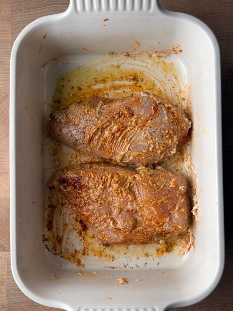 Raw chicken breasts coated in a lemon Greek yogurt marinade in a white rectangular baking dish.