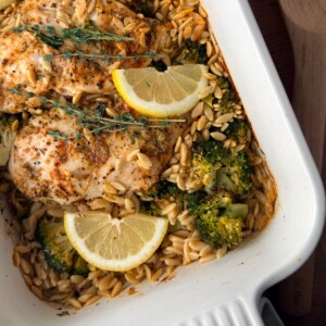 Lemon chicken orzo pasta bake with broccoli, fresh thyme and lemon wedges in a white baking dish.