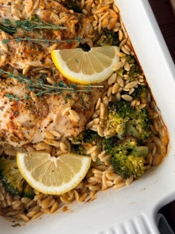 Lemon chicken orzo pasta bake with broccoli, fresh thyme and lemon wedges in a white baking dish.