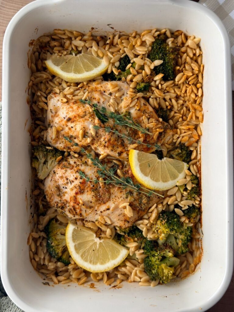 Finished lemon chicken orzo pasta bake with broccoli, fresh thyme and lemon wedges in a white baking dish.