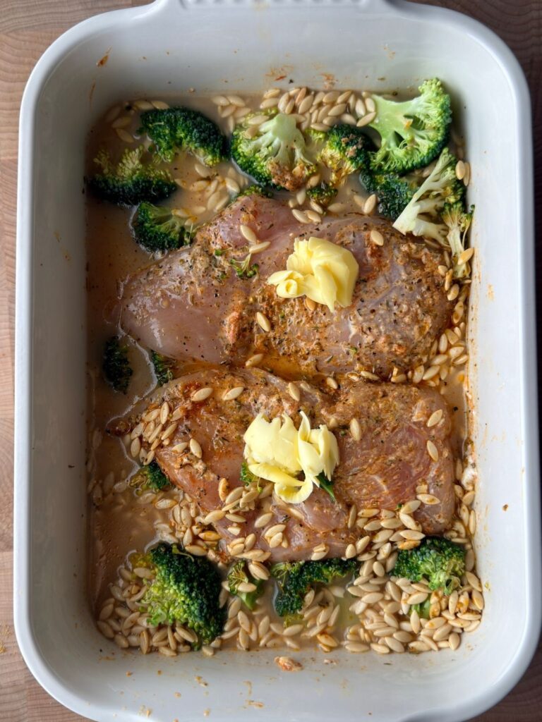 Lemon chicken orzo pasta bake before cooking, with chicken breasts, broccoli, orzo, water and butter in a white baking dish.