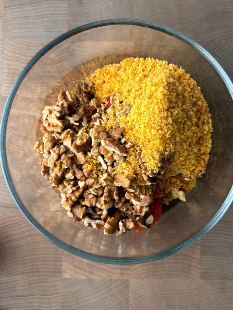Muhammara recipe ingredients in a food processor bowl before blending, including roasted red peppers, walnuts and toasted panko.