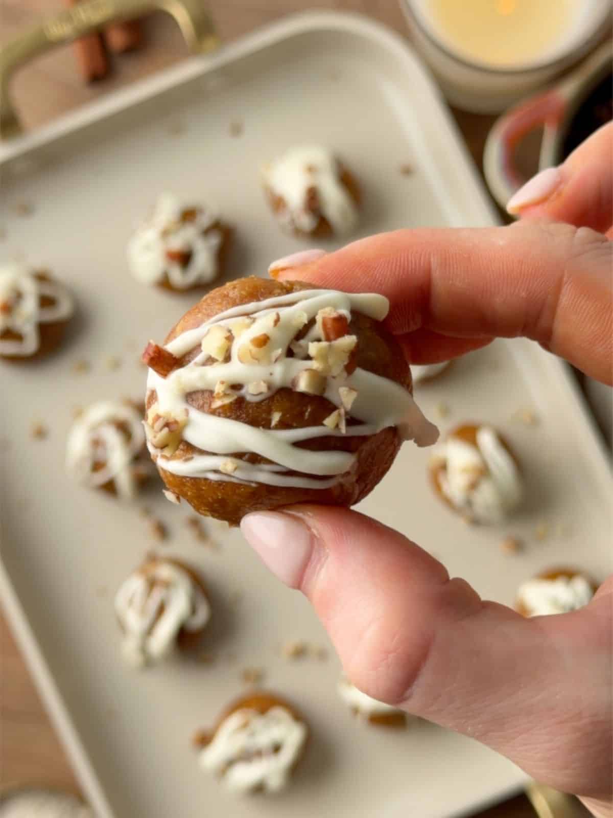 Pumpkin protein ball with white chocolate drizzle and chopped pecans held between two fingers, with more protein balls in the background.