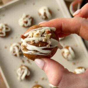 Pumpkin protein ball with white chocolate drizzle and chopped pecans held between two fingers, with more protein balls in the background.