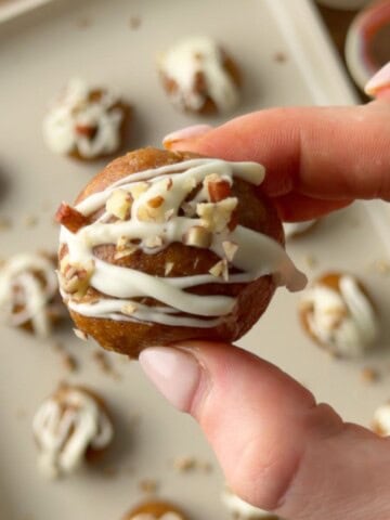 Pumpkin protein ball with white chocolate drizzle and chopped pecans held between two fingers, with more protein balls in the background.