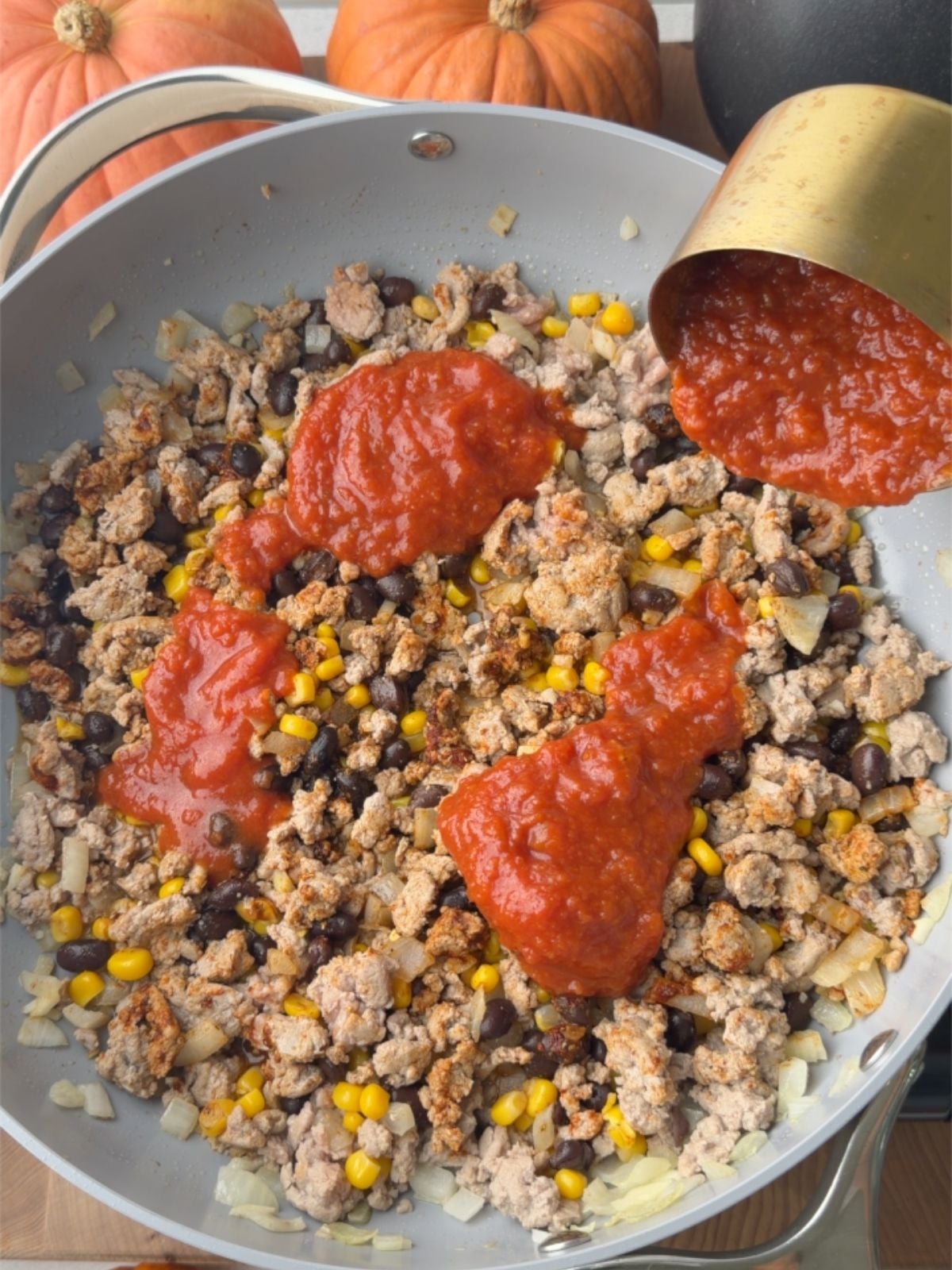 Pouring tomato sauce into a skillet of seasoned ground turkey, black beans, and corn for stuffed peppers.