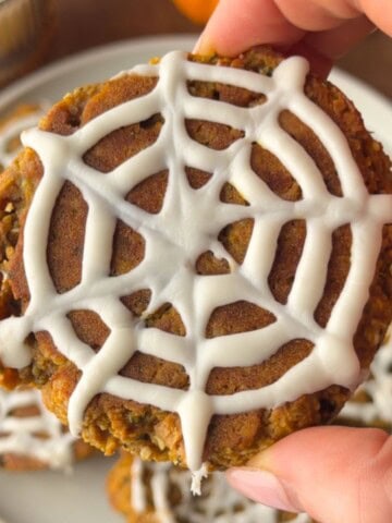 Soft pumpkin protein cookie topped with a white chocolate spiderweb design for an easy Halloween treat.