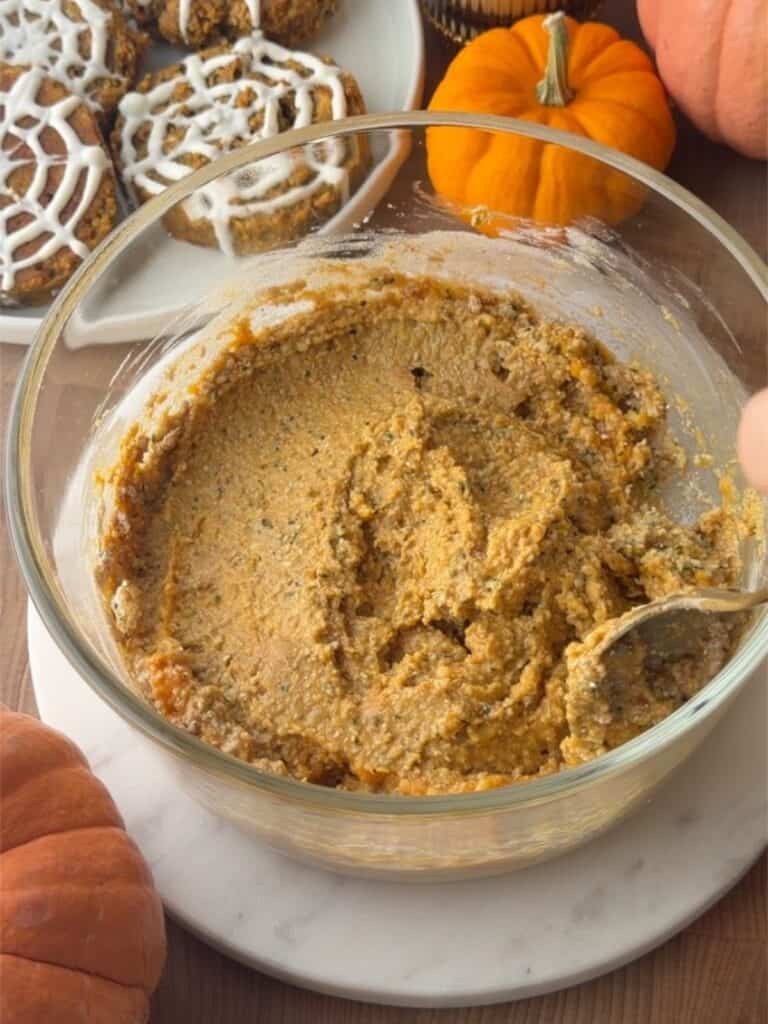 Pumpkin protein cookie dough mixed in a glass bowl, with finished Halloween cookies and mini pumpkins in the background.